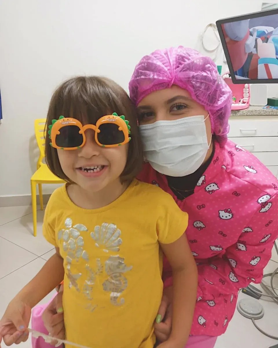 Uma menina de óculos de brinquedo em forma de dinossauro, abraçada pela dentista de touca rosa.