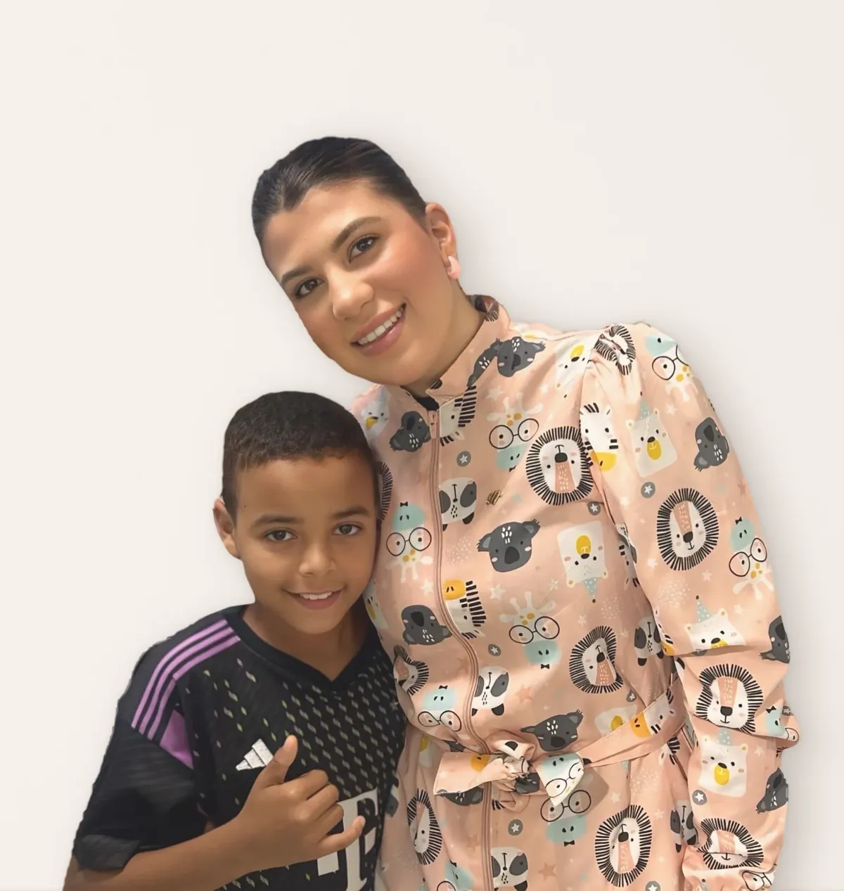 Uma dentista de jaleco estampado de bichinhos abraçada a um menino de camisa de futebol, que faz o sinal de hang loose com a mão.