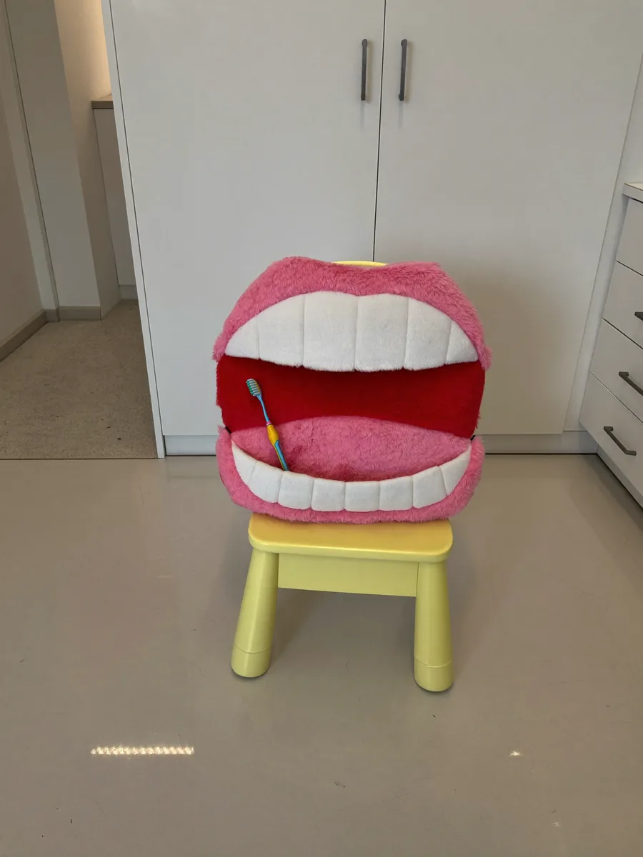 Uma pelúcia em forma de boca aberta, com dentes de feltro, ao lado de uma escova de dente infantil.