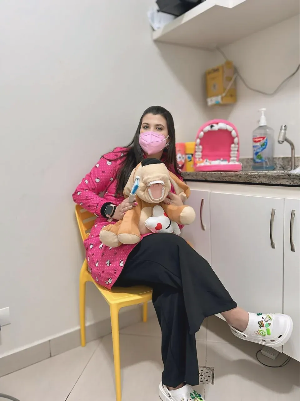 Dentista de jaleco rosa sentada numa cadeirinha amarela, com um cachorro de pelúcia de dentes à mostra no colo.